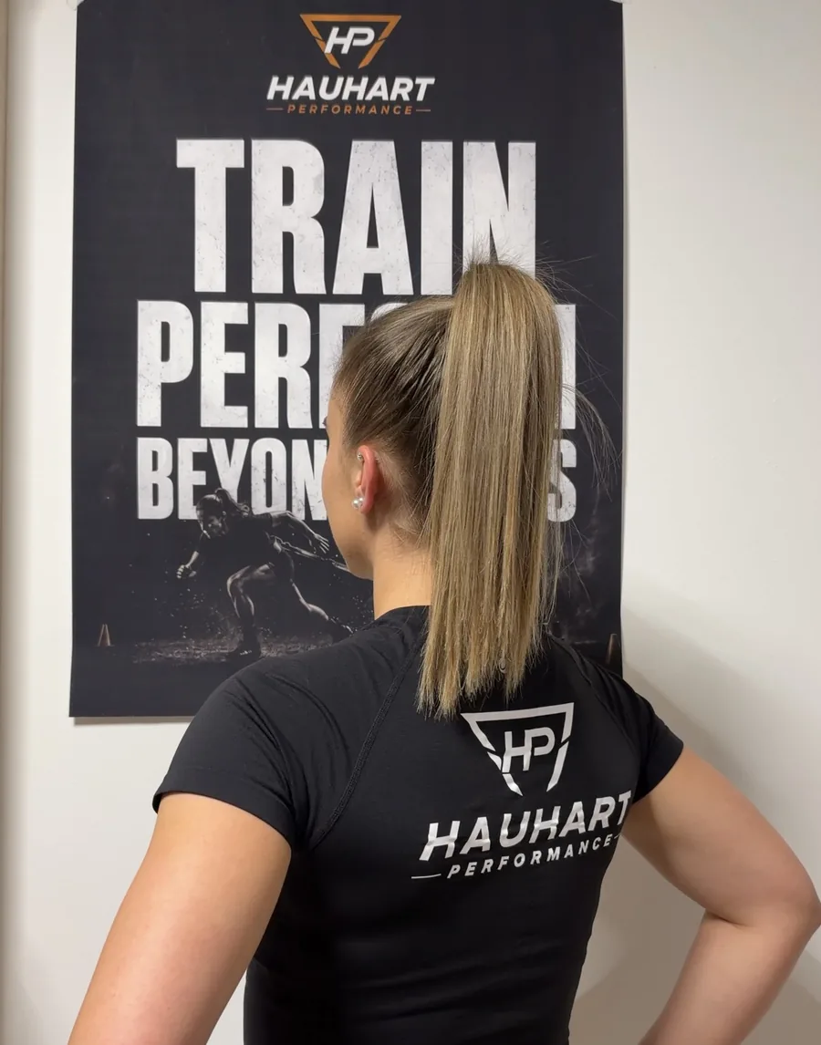 Johanna Hauhart von hinten im Hauhart-Performance-Shirt vor dem Train-Perform-Beyond-Plakat